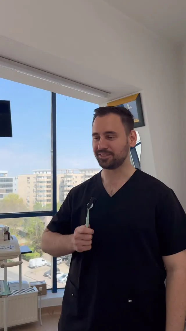 Dr. Radu Patriche, în halat negru, ținând o periuță de dinți lângă fereastra cabinetului.